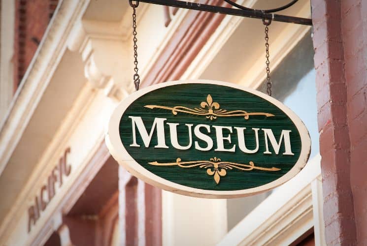 A green, gold and white oval museum sign hanging by a red and white building