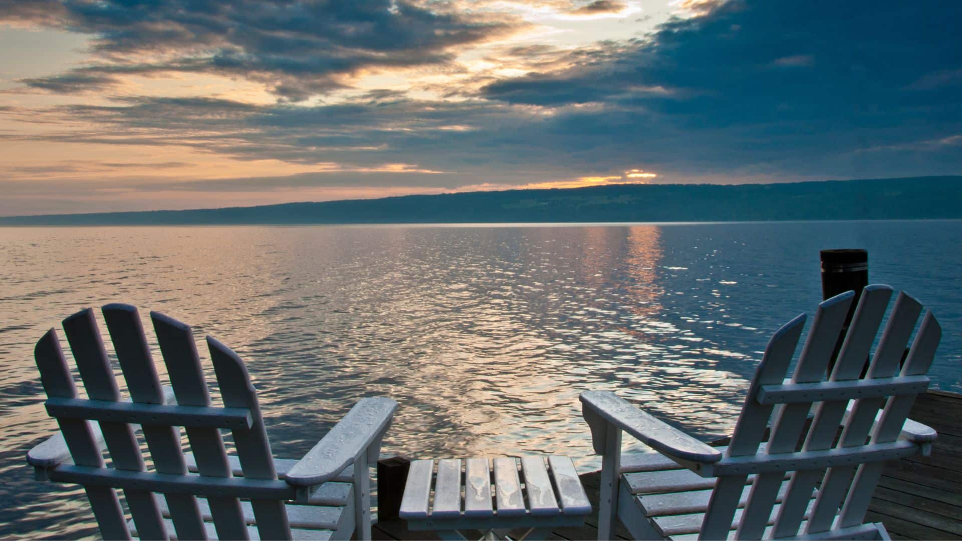 Two white Adirondack chairs at the end of a dock overlooking an expansive lake