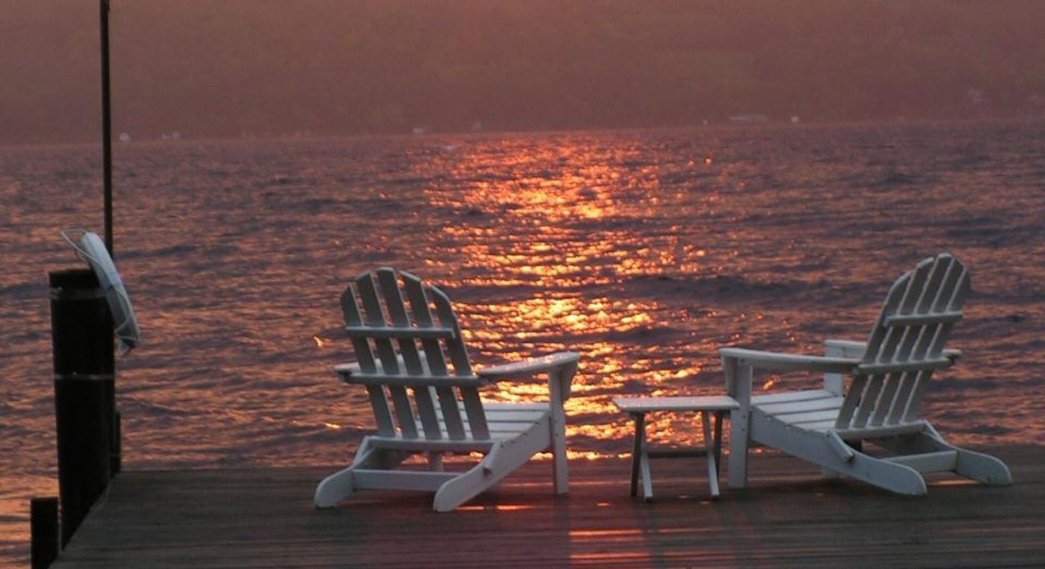 Two white Adirondack chairs at the end of a dock overlooking a large lake at sunset