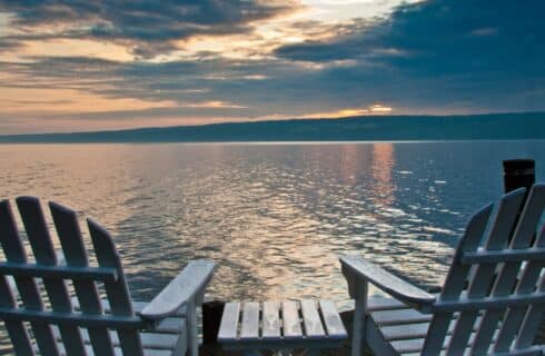 Two white Adirondack chairs and a side table at the end of a dock overlooking a large lake