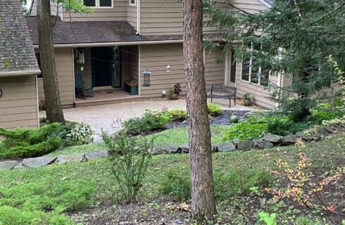 Front door of a large brown home nestled in the woods with lush green landscaping and tall trees