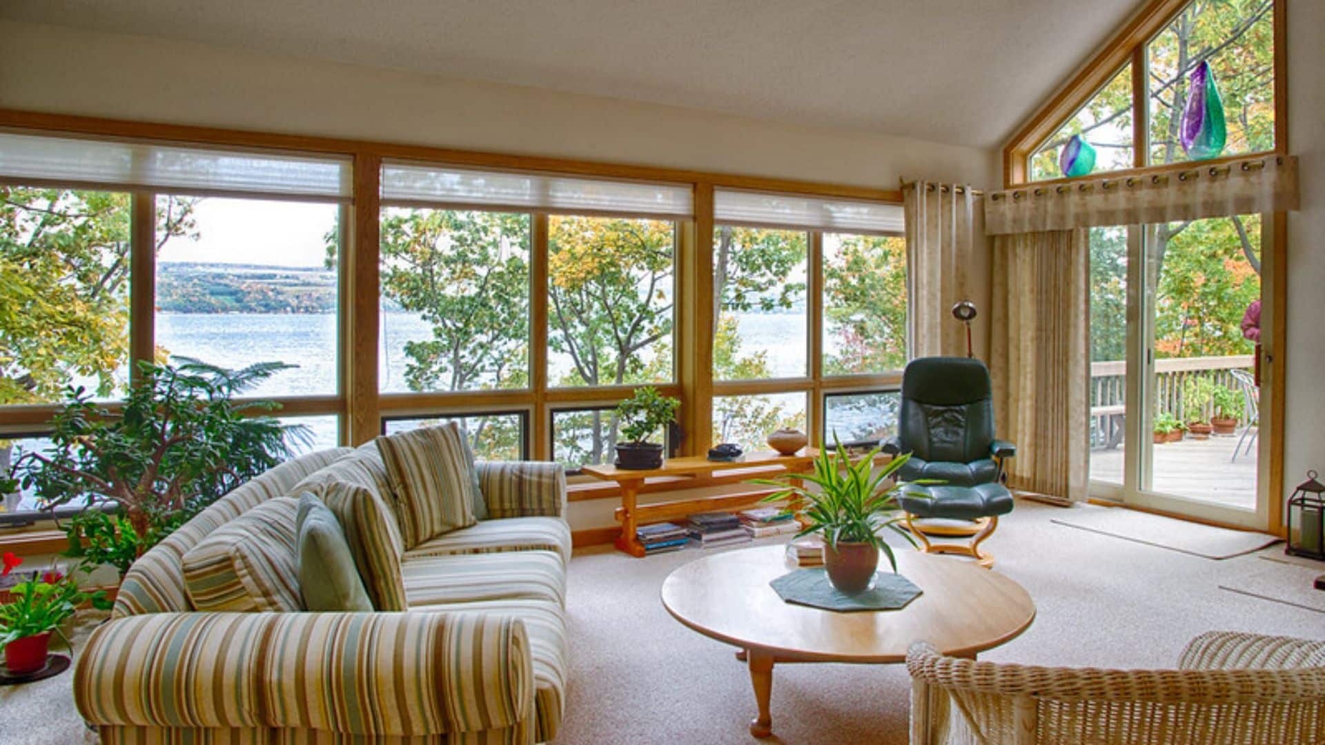 Spacious living room with couch, table and chairs and walls of windows overlooking a lake and trees