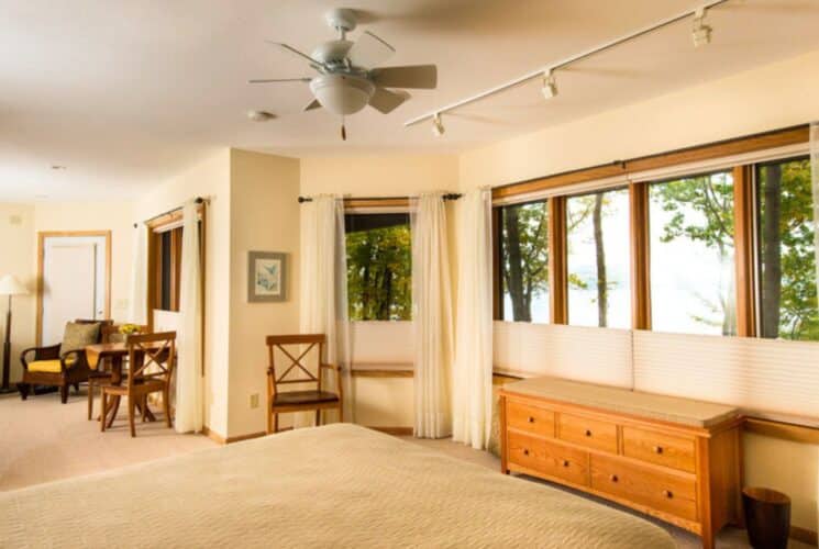 Spacious apartment suite with bed, couch, table and chairs, dresser and walls of windows overlooking a lake and trees