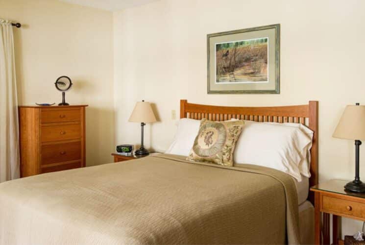 Bedroom with wood bed, cream and white linens, side tables with lamps and tall dresser next to a window with blinds and curtains