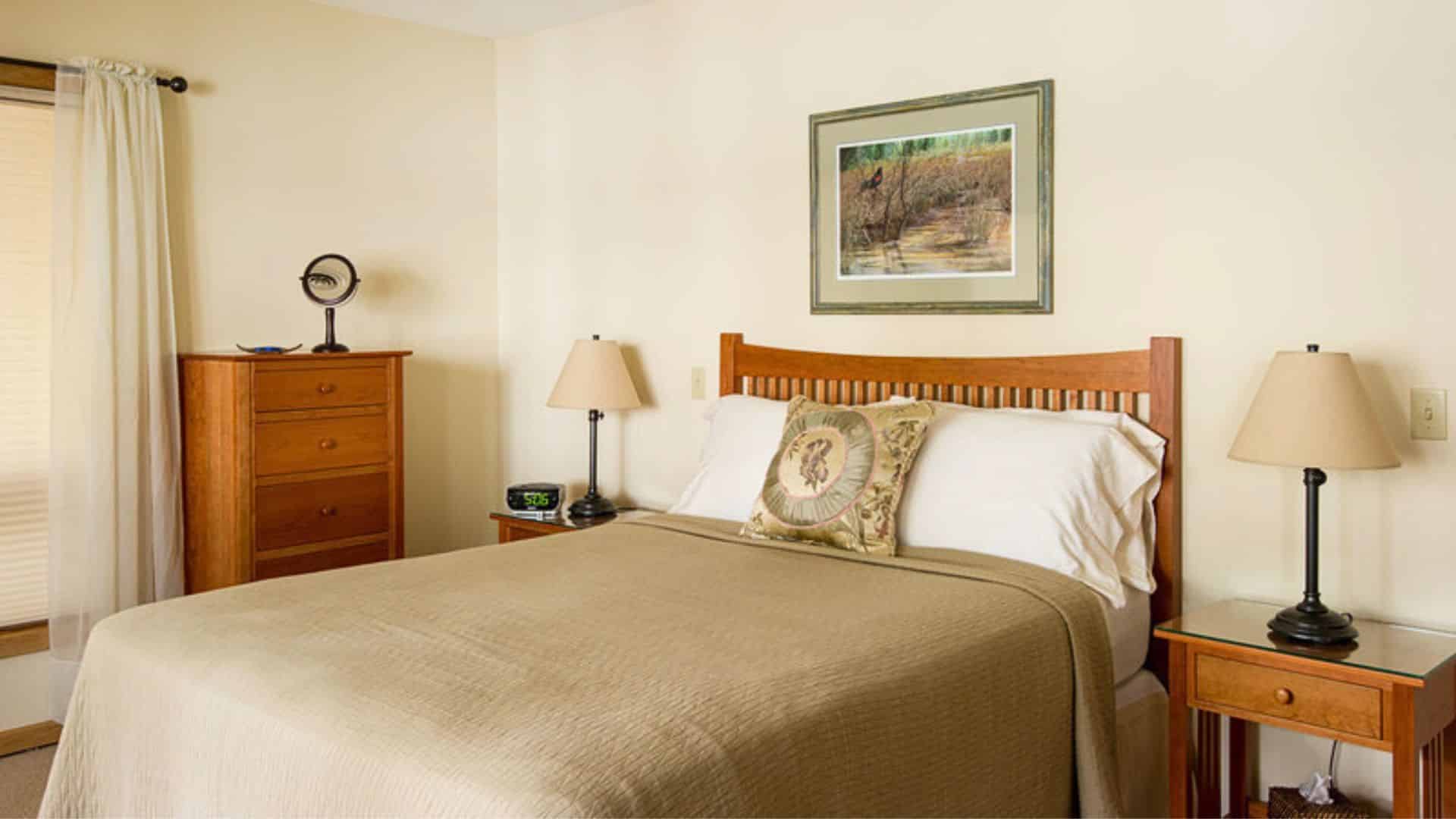 Bedroom with wood bed, cream and white linens, side tables with lamps and tall dresser next to a window with blinds and curtains