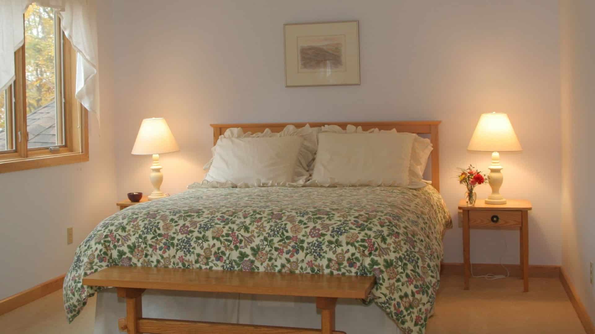 Bedroom with queen bed with a colorful floral quilt, side tables with lamps and window with white curtains