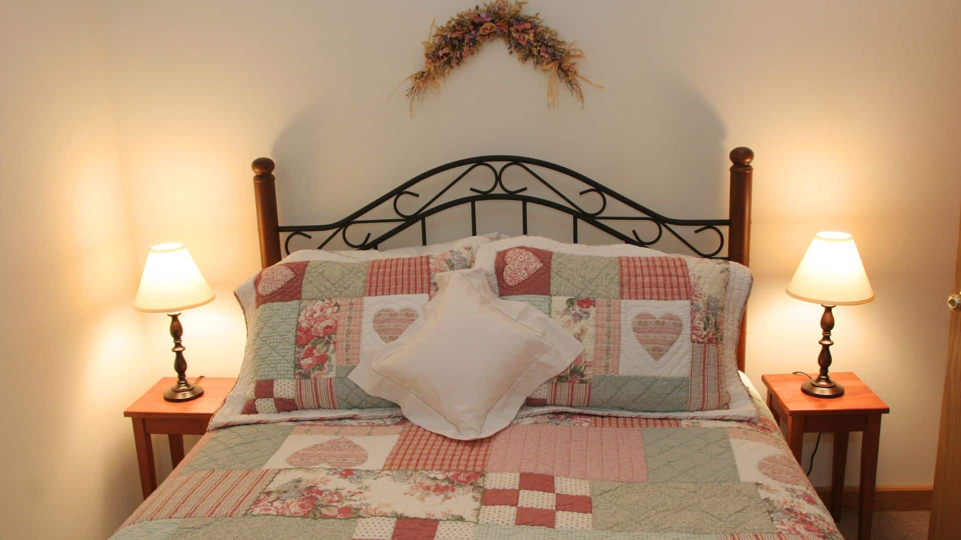 Bedroom with wood and iron bed with colorful patchwork quilt, side tables with lamps and floral swag over the headboard