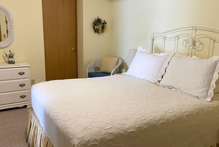 Bedroom with bed in white linens, white dresser, oval wicker mirror and white wicker sitting chair