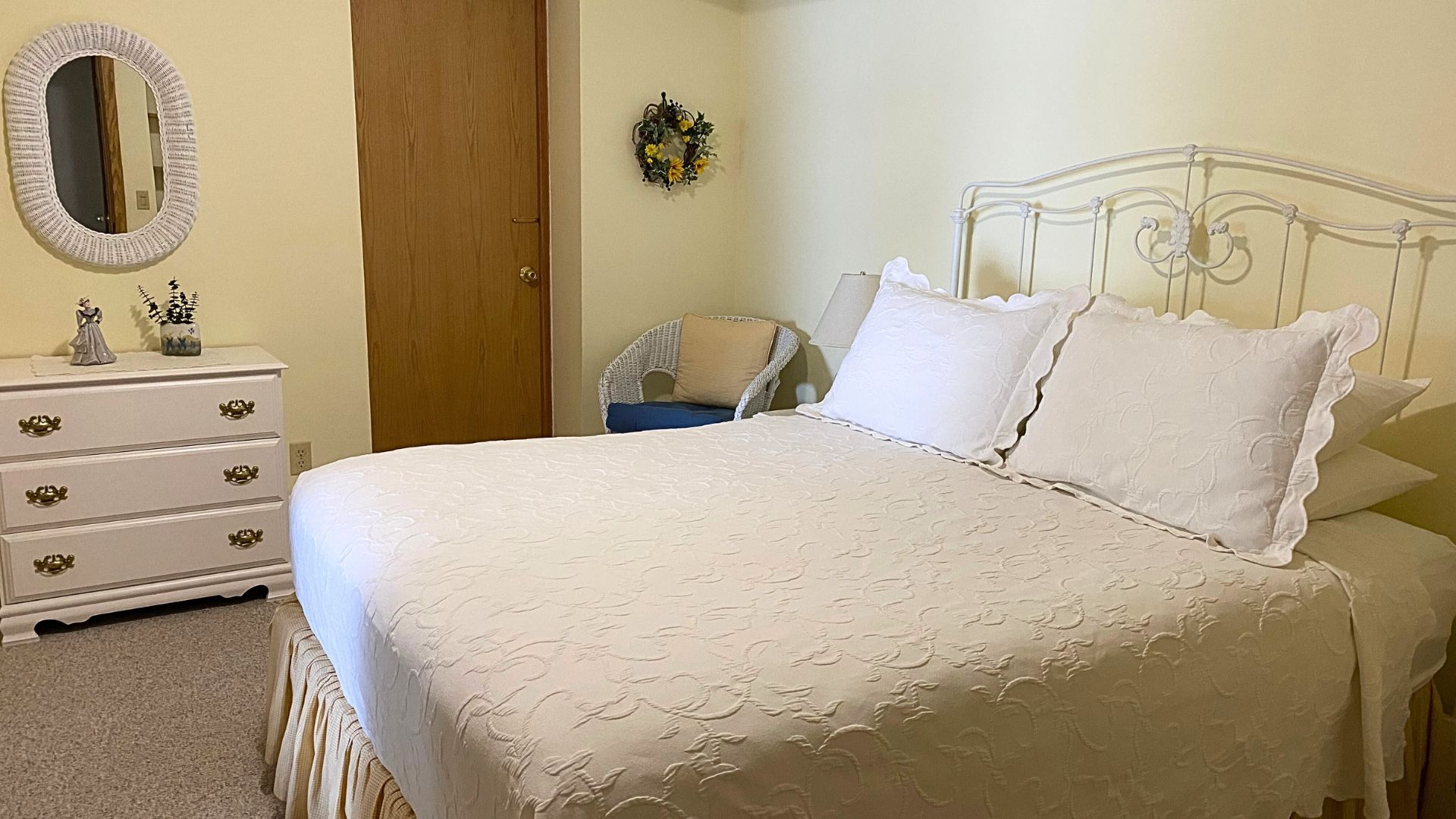 Bedroom with bed in white linens, white dresser, oval wicker mirror and white wicker sitting chair