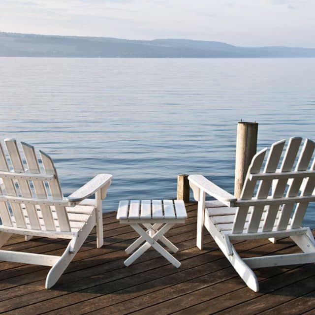 Two white Adirondack chairs on the edge of a dock by a large lake