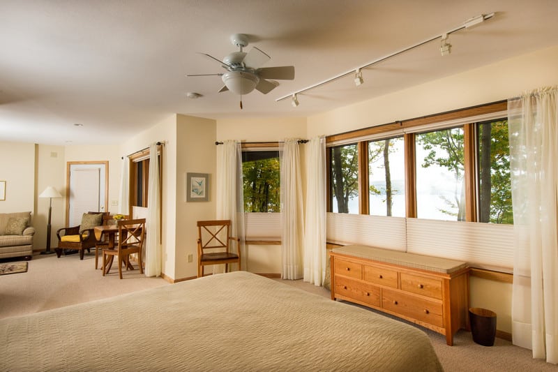 A spacious apartment suite with bed, couch, table and chairs and large windows overlooking trees and a lake