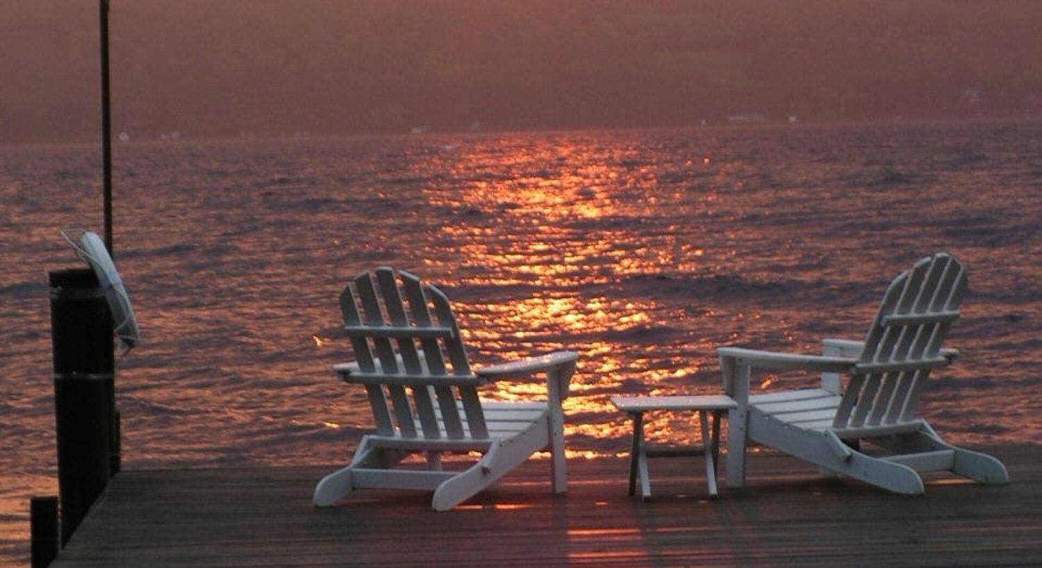 Two white Adirondack chairs at the end of a dock overlooking a large lake at sunset