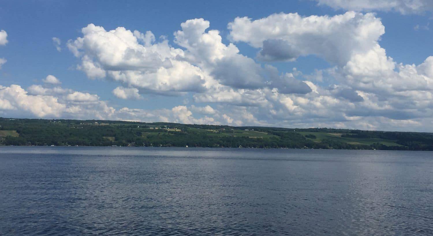 An expansive lake with tree covered hills at one end and clouds and blue skies above